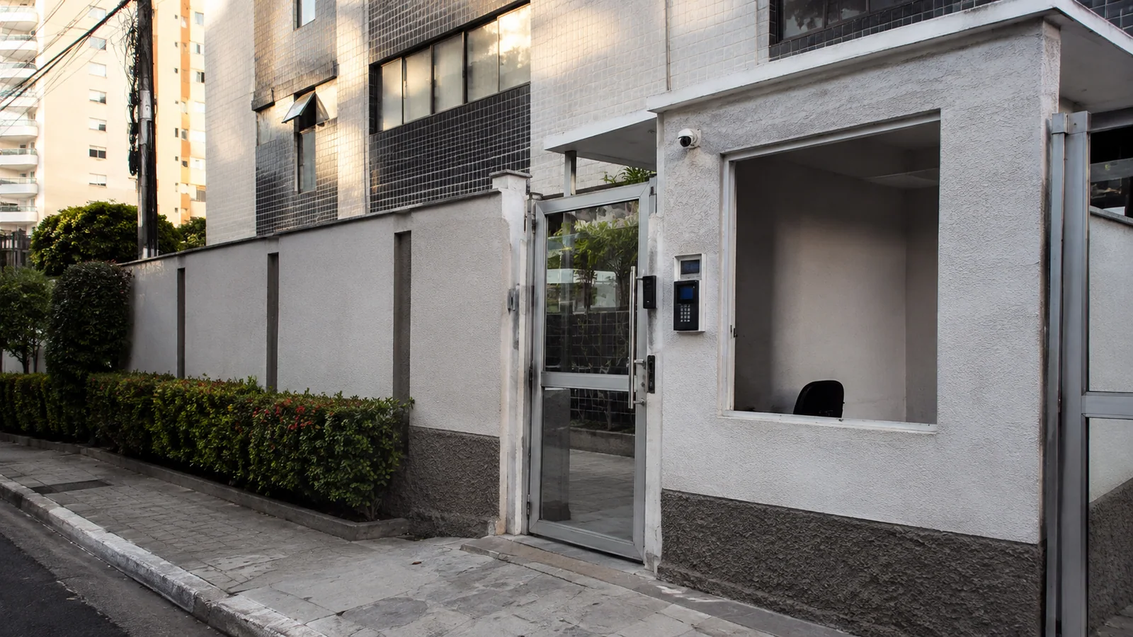 Entrada urbana de condomínio pequeno com terminal de acesso, interfone, câmera e guarita vazia