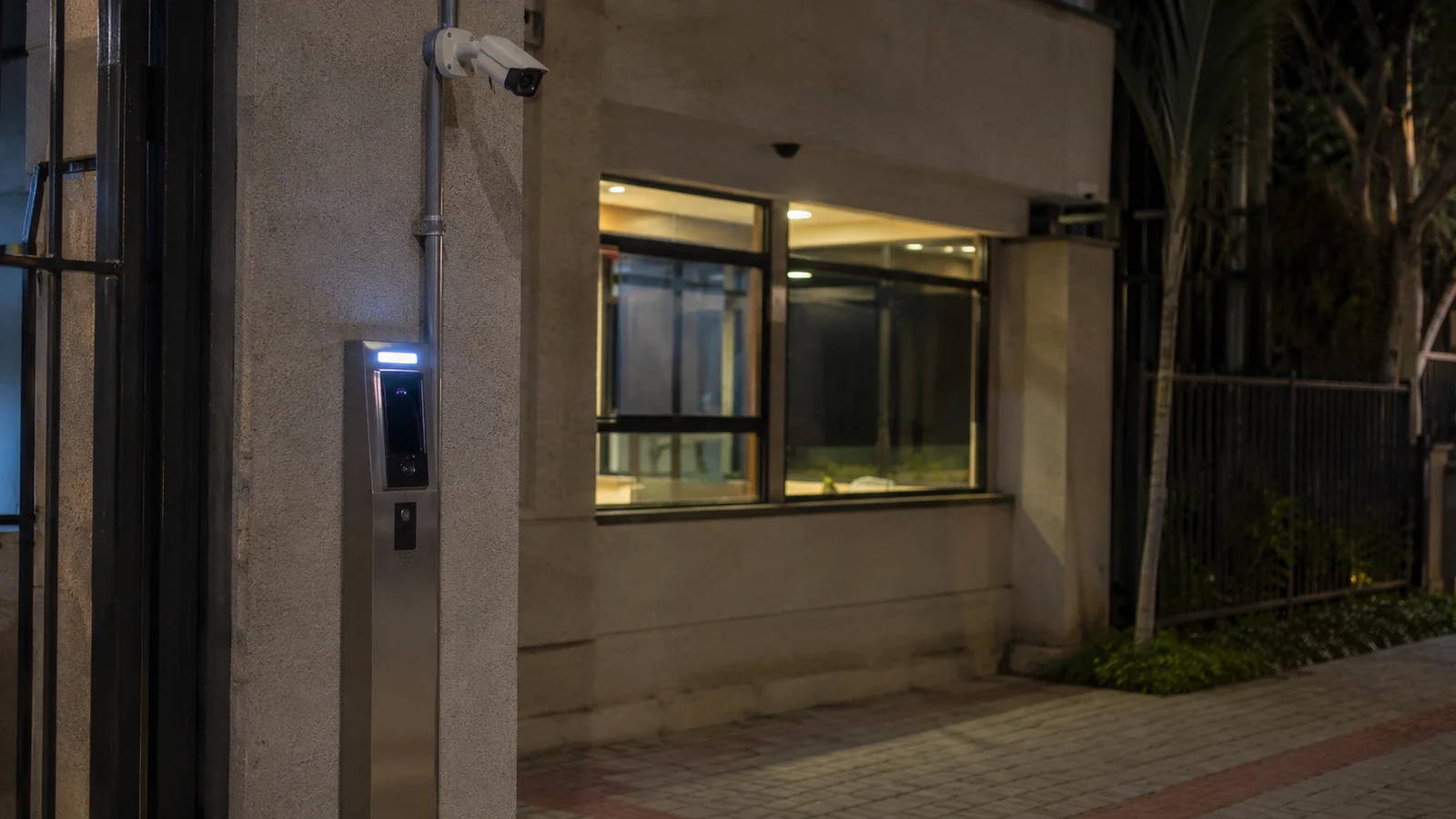 Entrada de pedestres de condomínio à noite com totem de acesso e terminal facial na coluna, câmera acima e guarita iluminada e vazia ao fundo